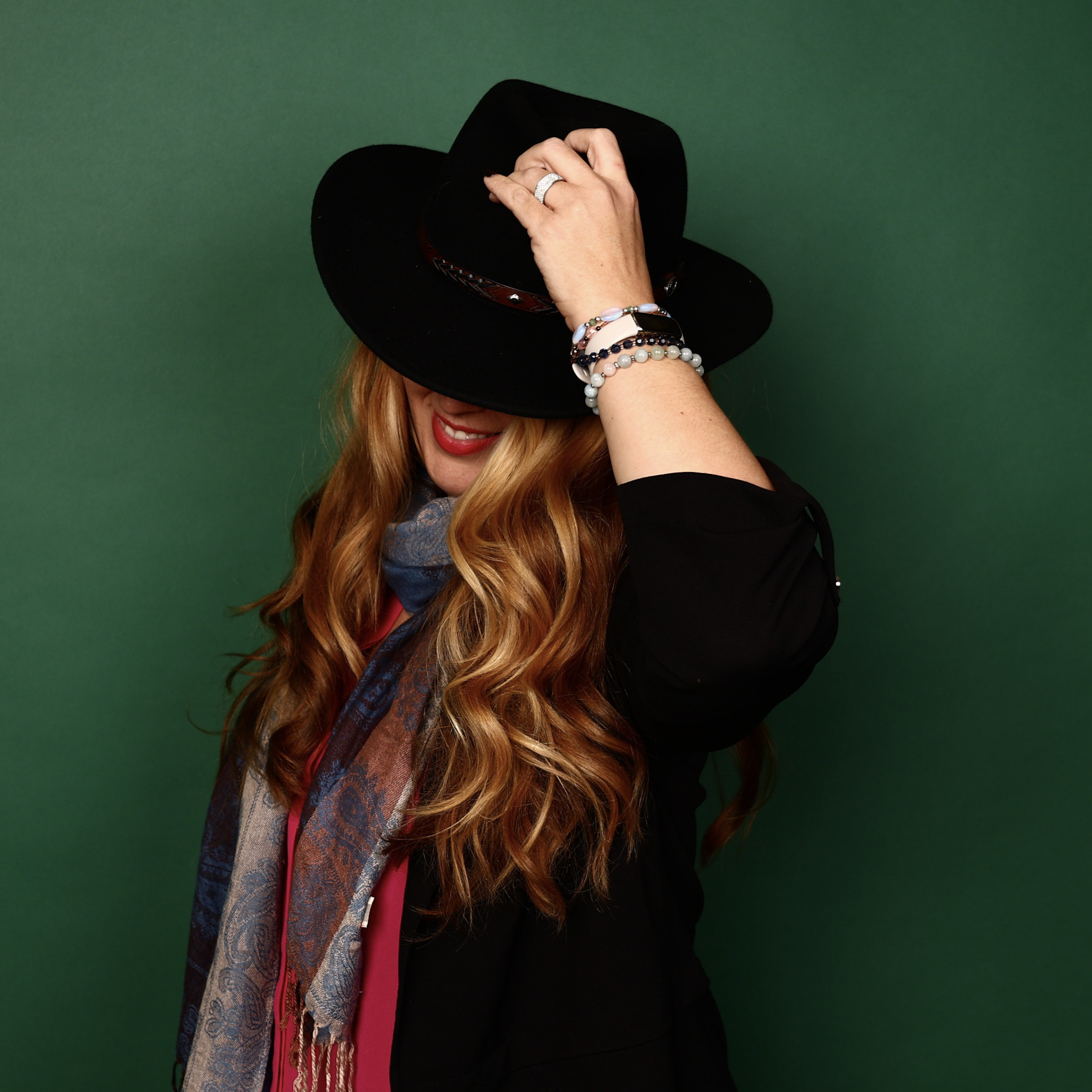 Upcoming Guest Jules Kuroda, a woman with long, wavy red hair, wears a black hat and scarf, smiling as her face is partially covered by the hat against a green background.